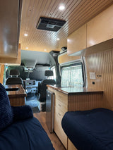 Spacious custom RV rental interior with wood paneling, recessed lighting, kitchen area, and comfortable seating, extending to the driver's cab.