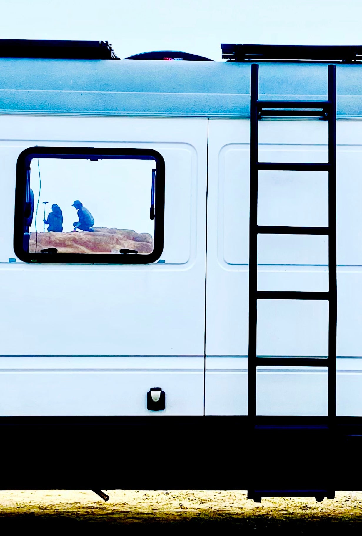 RV rental side view: A white camper van with a ladder. Its window frames two silhouetted figures on a miniature rocky scene.