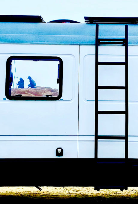 RV rental side view: A white camper van with a ladder. Its window frames two silhouetted figures on a miniature rocky scene.
