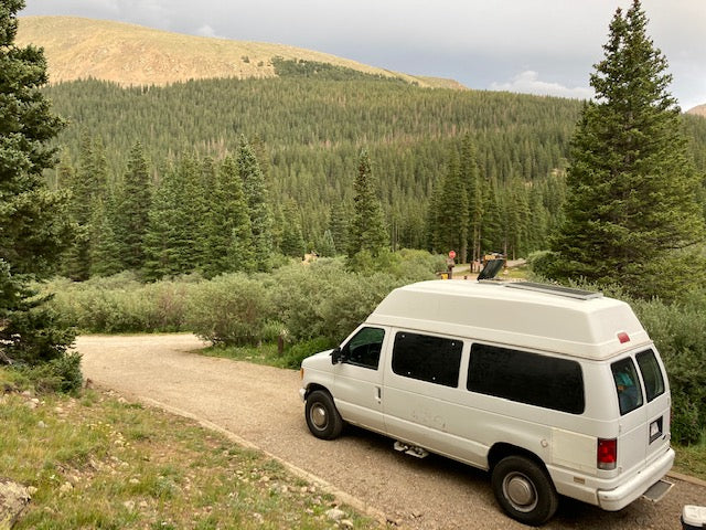 An RV rental van, white with a high top and solar panels, parked on a gravel path in a dense mountain forest.