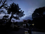 An RV rental with a rooftop tent parked at a dark campsite under a cloudy night sky, surrounded by large silhouetted trees.