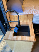 RV rental's compact kitchen: a black faucet and sink set in a wood countertop, with a blue sponge, and a cozy bed visible.