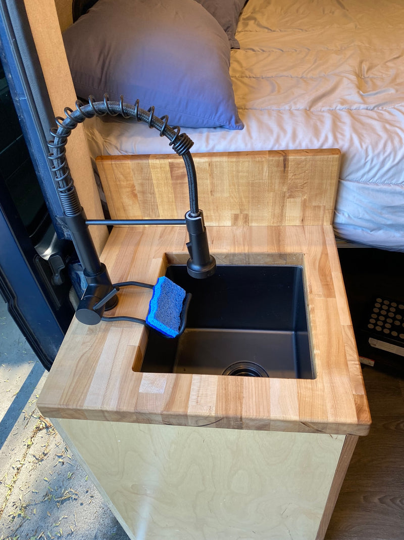 RV rental's compact kitchen: a black faucet and sink set in a wood countertop, with a blue sponge, and a cozy bed visible. — photo 9
