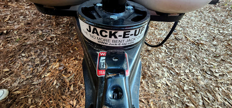 "JACK-E-UP" device on an RV rental trailer hitch, preventing bent jacks. Ground covered in pine needles and leaves.