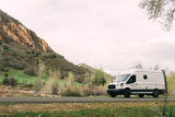 White RV rental van parked on a mountain road with lush green hills and distant snow-capped peaks under a cloudy sky.