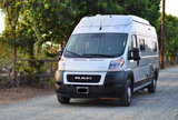 A modern white Ram camper van, ideal for an RV rental road trip, parked on a sunlit gravel path surrounded by trees.