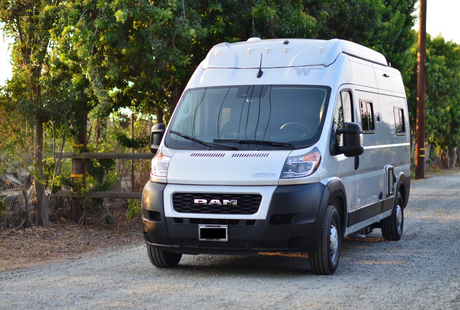 A modern white Ram camper van, ideal for an RV rental road trip, parked on a sunlit gravel path surrounded by trees.