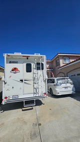 A white rv rental with a Jurassic Park decal is parked in a driveway next to a minivan and a house on a clear day.