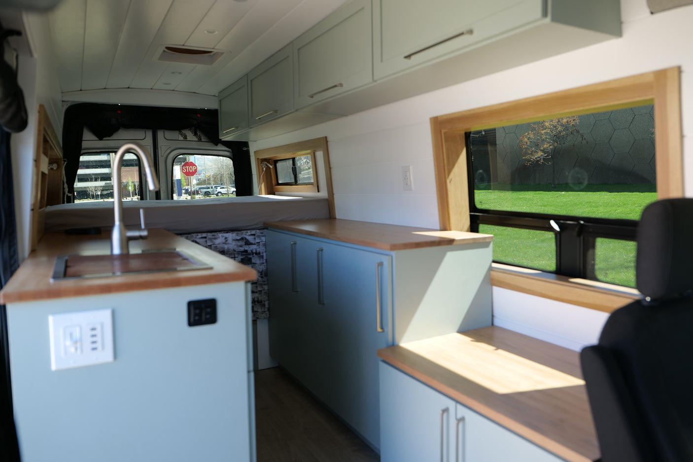 Step inside this modern RV rental van conversion featuring a custom kitchen with light blue cabinets, wood counters, and a cozy bed.