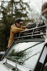 A smiling man in a yellow jacket secures a case onto a vehicle's roof rack with a friend. Prepping for an outdoor adventure, like those enjoyed with an rv rental.