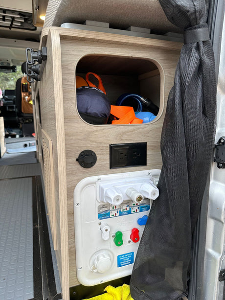 RV rental's compact interior: water utility panel with hookups, power outlet, and storage cubby.