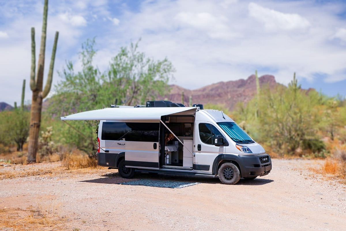 A white camper van RV rental with an extended awning is parked on a dirt road in a sunny desert landscape