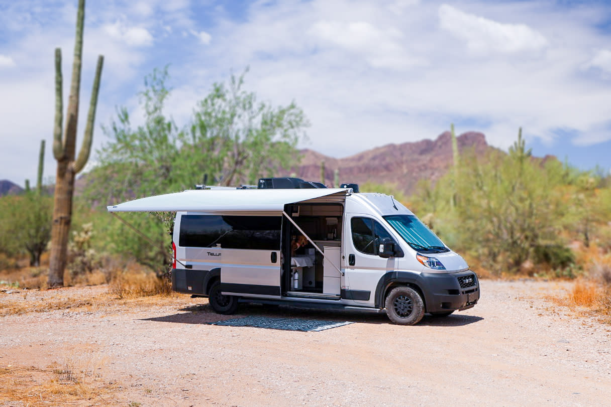 A white camper van RV rental with an extended awning is parked on a dirt road in a sunny desert landscape, ideal for outdoor living.