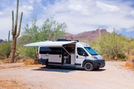 A white camper van RV rental with an extended awning is parked on a dirt road in a sunny desert landscape, ideal for outdoor living.
