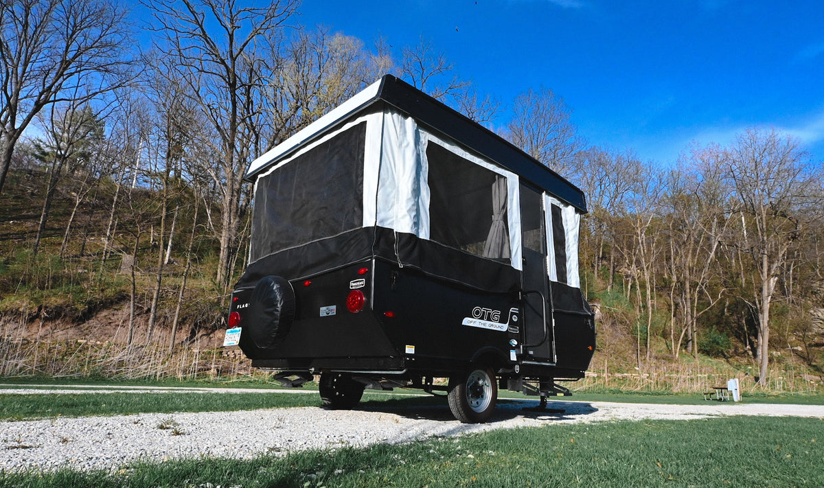 This pop-up RV rental is set up on a gravel path at a wooded campsite, ready for adventure under a clear blue sky.