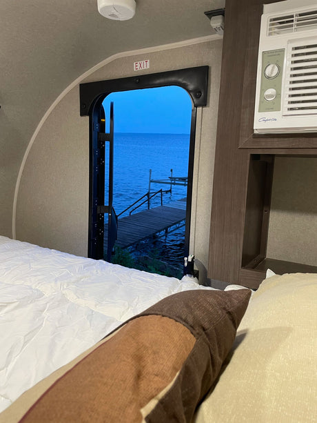 Experience a peaceful rv rental escape! Cozy bed inside a travel trailer with open door view of a calm blue lake & private dock.