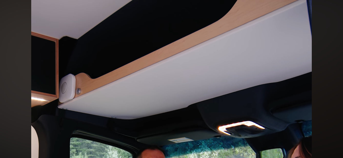 RV rental interior: Overhead storage with wood trim, a white sensor, and the illuminated front cabin. Trees through the windshield.