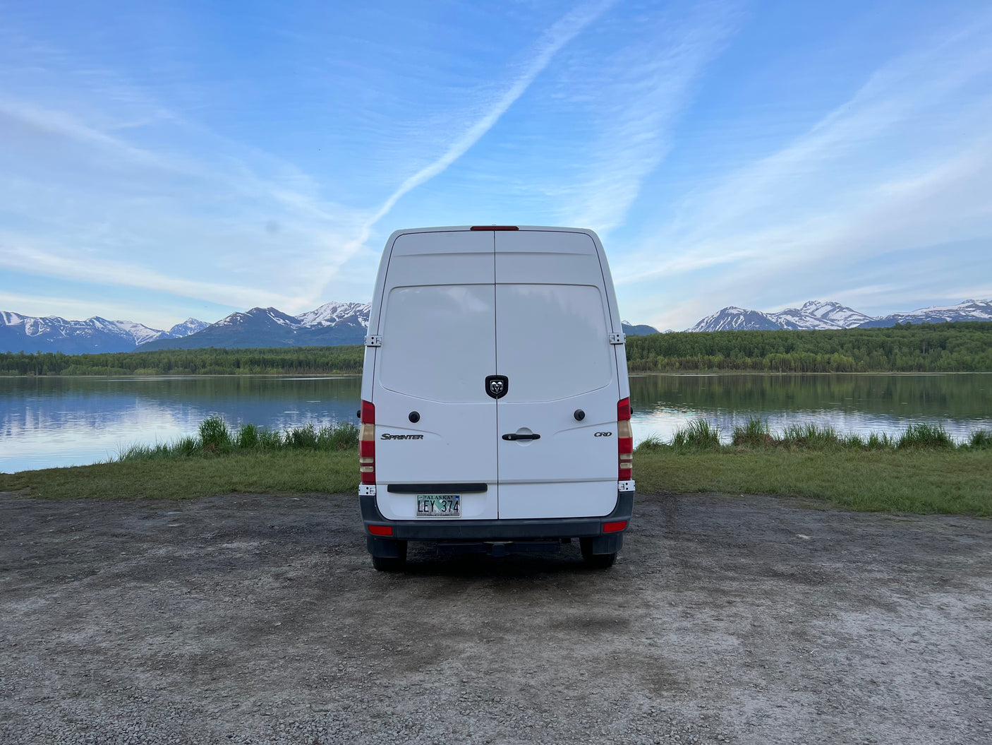 A white Sprinter van (RV rental) by an Alaskan lake, with mountains & forests. Ready for adventure!
