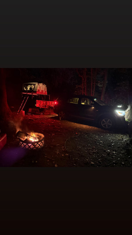 Night adventure camping with an RV rental alternative: a rooftop tent on a trailer, cozy campfire, and SUV under trees.