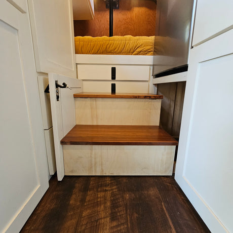 Cozy RV rental interior: Wooden steps lead to a yellow-quilted bed with storage, white cabinetry, and dark wood floors.