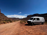 Adventure-ready RV rental Sprinter van on a red dirt road in a vast desert canyon landscape under a blue sky.