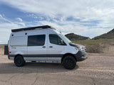 A customized silver Sprinter camper van (rv rental) on a gravel road in a desert. Features an awning & off-road tires.
