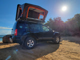 A black SUV with an open rooftop tent is parked on a sunny beach next to the ocean, perfect for an RV rental adventure.
