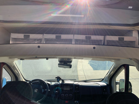 View from inside an RV rental: driver's area, dashboard, windshield, and overhead storage bins. Sunlight flares.