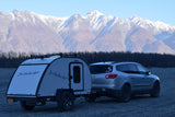 Bushwhacker RV rental and SUV on gravel, against majestic snow-capped mountains and bare trees.