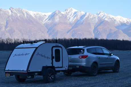 Bushwhacker RV rental and SUV on gravel, against majestic snow-capped mountains and bare trees.