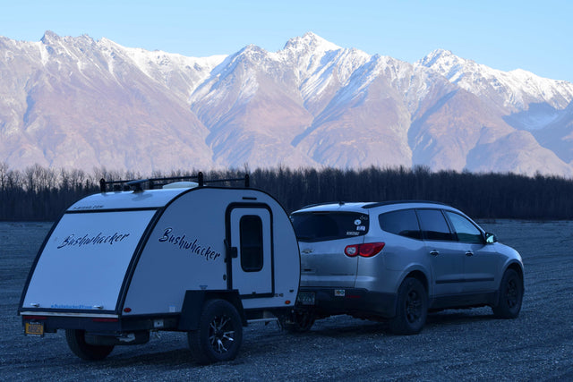 Bushwhacker RV rental and SUV on gravel, against majestic snow-capped mountains and bare trees.