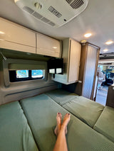 Comfy rv rental interior: feet on the green bed, TV, window view, and storage. Ideal for relaxing camper travel.