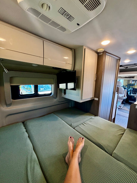 Comfy rv rental interior: feet on the green bed, TV, window view, and storage. Ideal for relaxing camper travel.