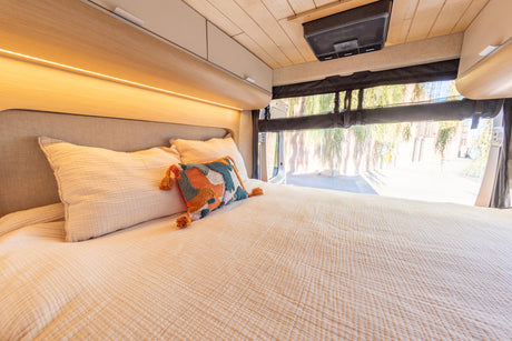 Comfortable RV rental interior featuring a cozy bed with ambient lighting, overhead storage, and outdoor views through the rear.