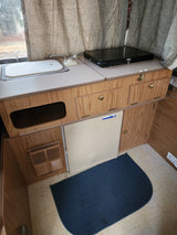 RV rental kitchenette: sink, covered stovetop, compact Norcold refrigerator, wood cabinets, and floor mat visible.
