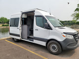 A white rv rental camper van with its side door open, revealing the interior layout, parked by a scenic river.