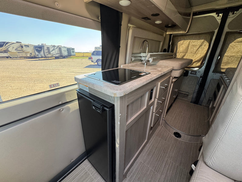 Modern rv rental interior: kitchen with sink, cooktop, and fridge, beside a window revealing parked RVs in an outdoor lot. — photo 6
