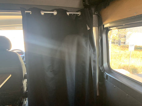 Interior of an RV rental campervan with a black privacy curtain separating the front cab from the living area, and an outdoor view.