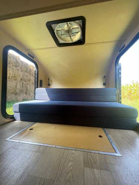 This RV rental offers a compact interior with a comfy convertible bed, ceiling fan, and windows showcasing rural views.