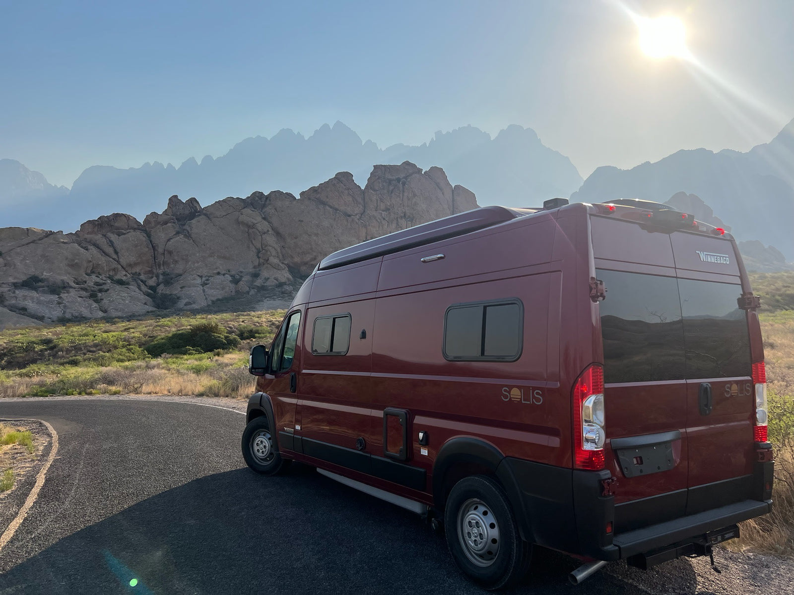 Red Winnebago Solis rv rental on a winding road, mountains in background, sun blazing. Ideal for a scenic road trip.