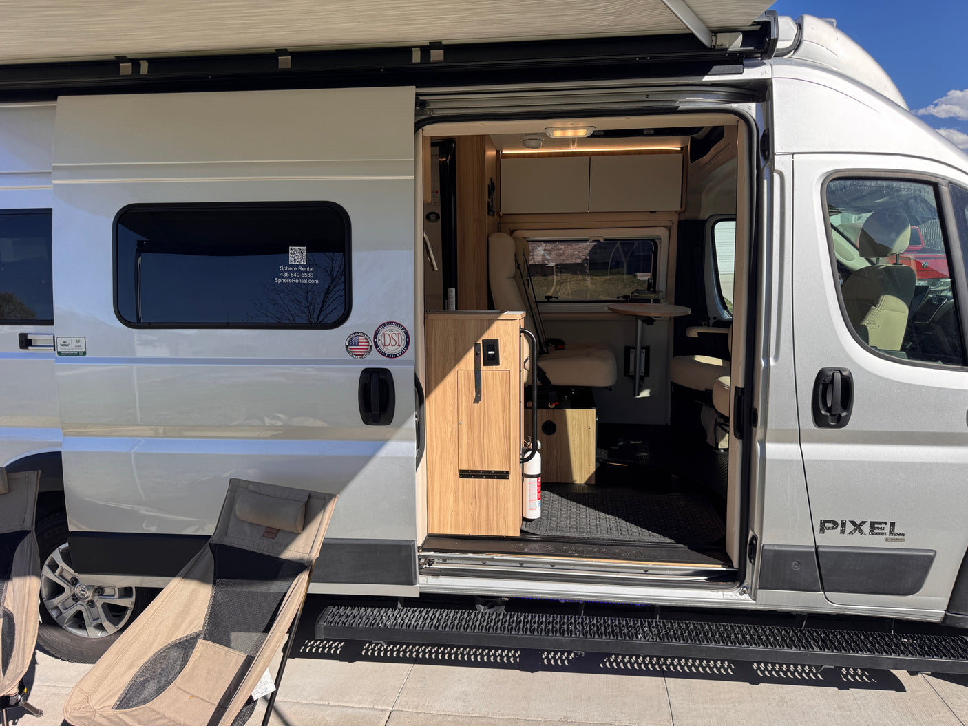 Open side door of a silver Pixel camper van rv rental, showcasing its interior, extended awning, and outdoor chairs.