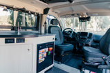 RV rental interior: light wood kitchenette with sink, fridge, and decorative mountain garland. Driver's cabin in view.