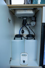 RV rental under-sink cabinet showing compact water system with two tanks, pump, and plumbing for a mobile kitchen.