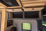 Interior of an RV rental: open wooden overhead cabinets, a control panel, and a side window view of green grass.