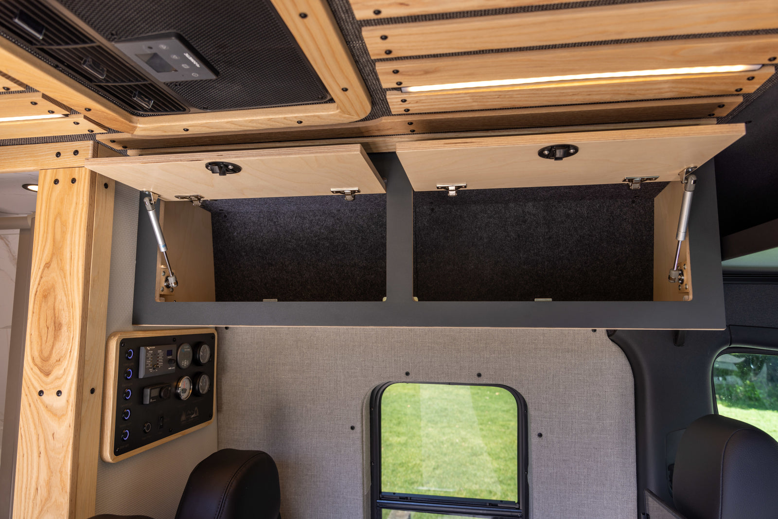 Interior of an RV rental: open wooden overhead cabinets, a control panel, and a side window view of green grass.