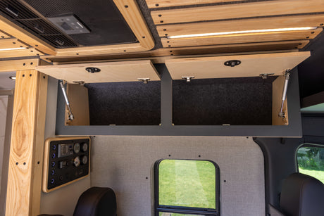 Interior of an RV rental: open wooden overhead cabinets, a control panel, and a side window view of green grass.