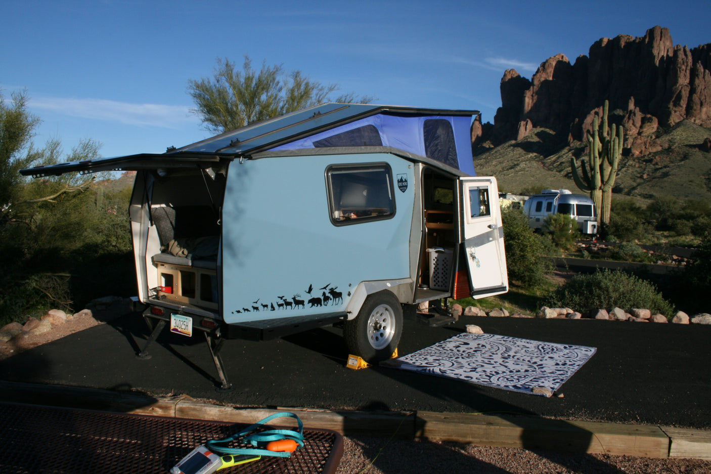 Enjoy desert adventures! This compact teardrop RV rental, with its pop-top tent, offers cozy camping amidst mountains & cacti.