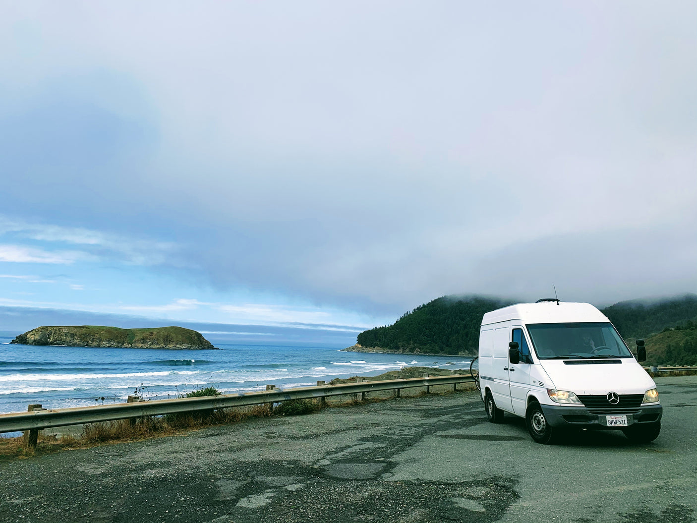 White Sprinter van on a scenic coastal overlook with ocean waves and an island, ideal for an RV rental adventure.