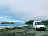White Sprinter van on a scenic coastal overlook with ocean waves and an island, ideal for an RV rental adventure.