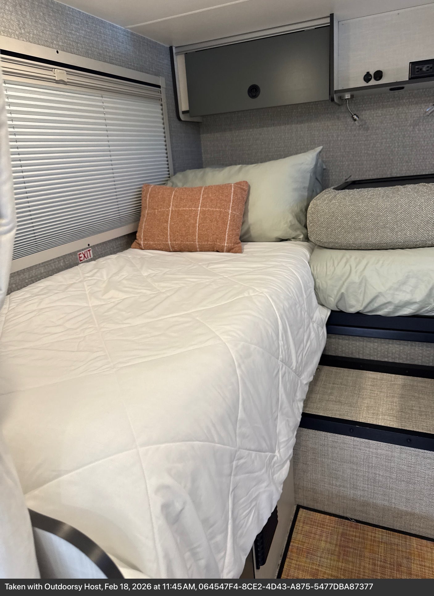 Cozy RV rental interior featuring a comfortable bed with white bedding, pillows, window blinds, and overhead storage.